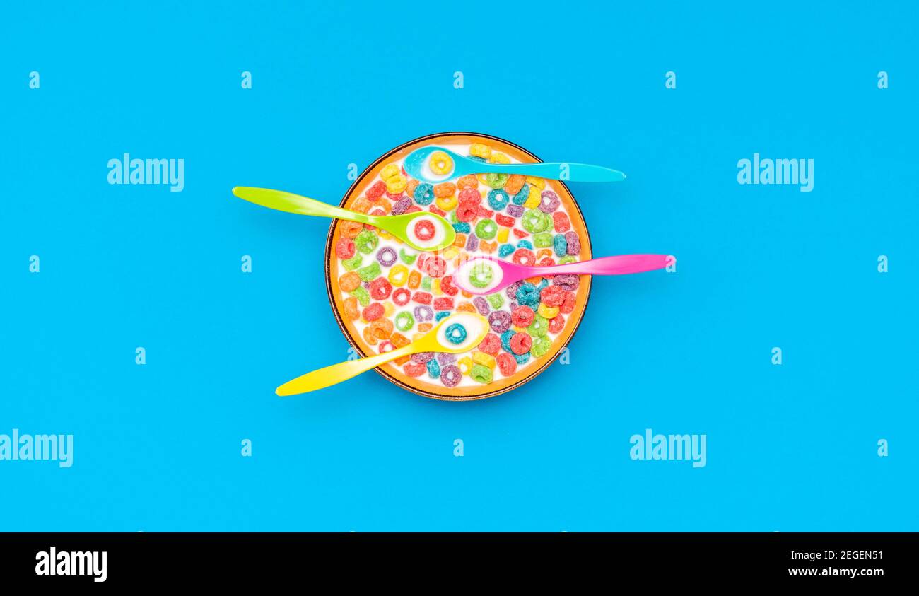 Top view with multicolored fruit rings cereals with milk in a bowl. Cereal bowl with four multicolored spoons isolated on a colored background. Stock Photo