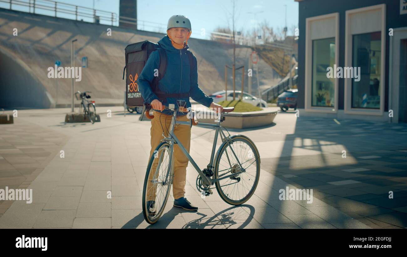 Handsome Happy Food Delivery Man Wearing Thermal Backpack and Safety Helmet Stands Beside his Bike in the Stylish Modern City District. Portrait of Stock Photo