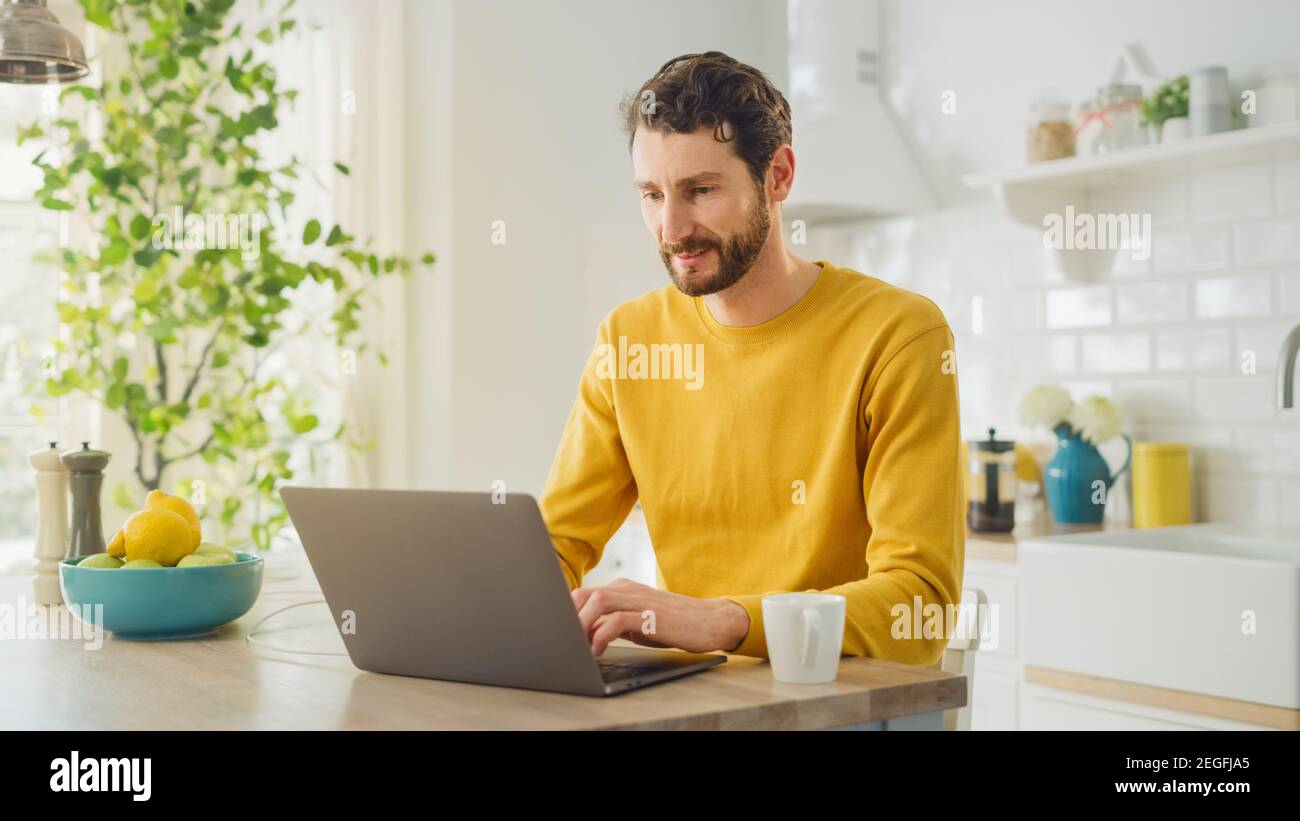 Young Handsome Man Using Laptop Computer and Drinking Morning Cup of Coffee or Tea While Wearing a Yellow Pullover. Curly-hared Male Sitting in a Stock Photo