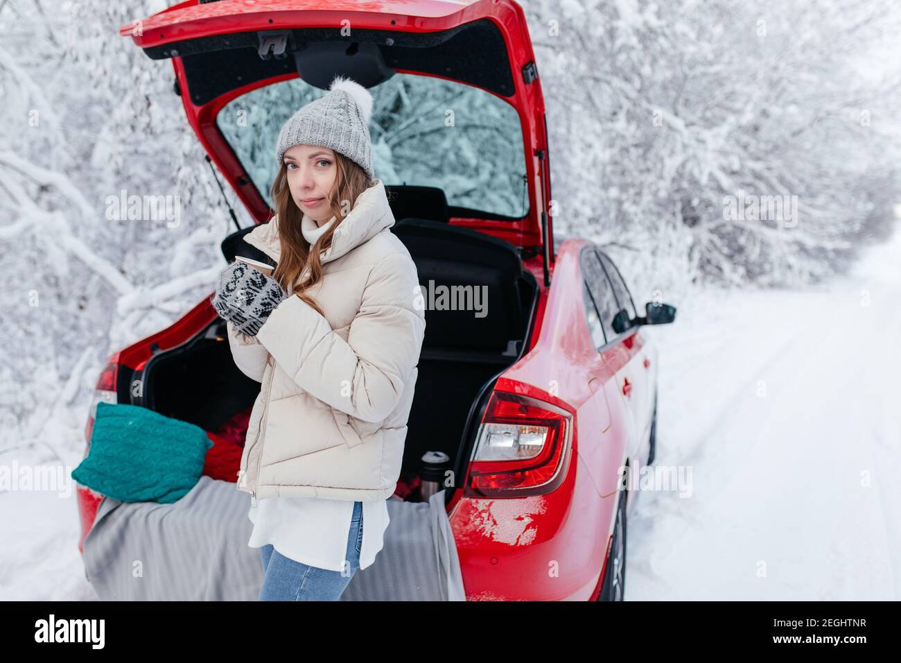 A woman with hot coffee in her hands sits in a red car on a snowy winter  day in the forest. A girl with a cup of hot coffee is sitting in
