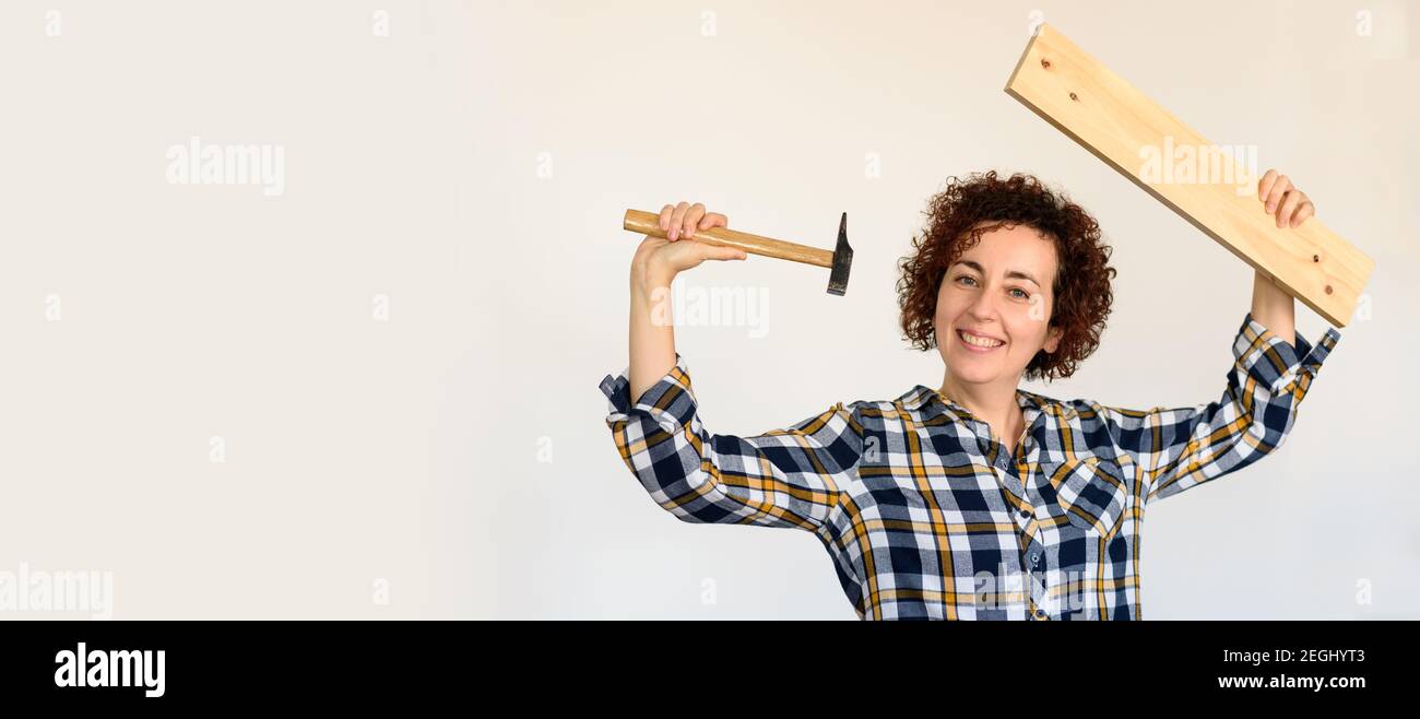 Young curly-haired caucasian girl holds a wooden board and a hammer. She raises her arms to show him how to camp. Interior of a flat. She white wall. Stock Photo
