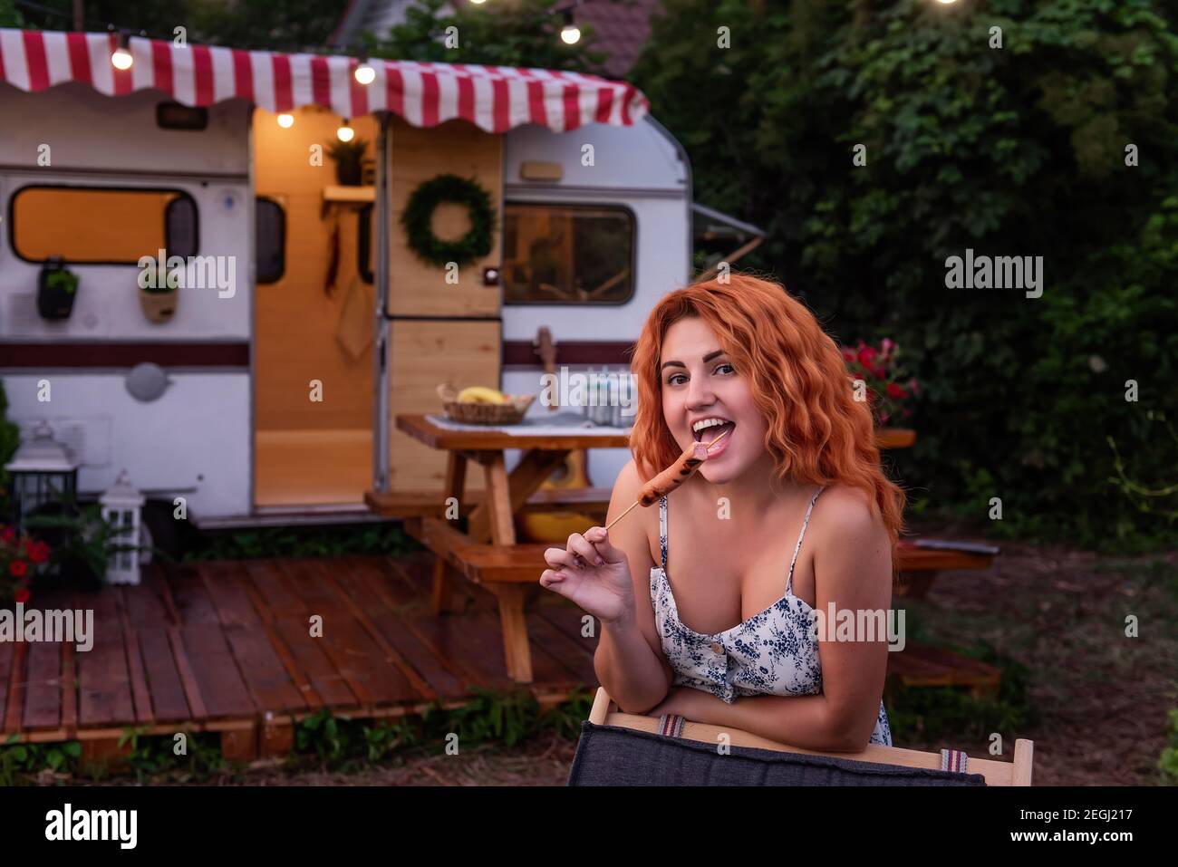 A young red-haired girl holds a barbecue sausage in her hands near the trailer track. The girl spends her vacation with her family in the country, hav Stock Photo