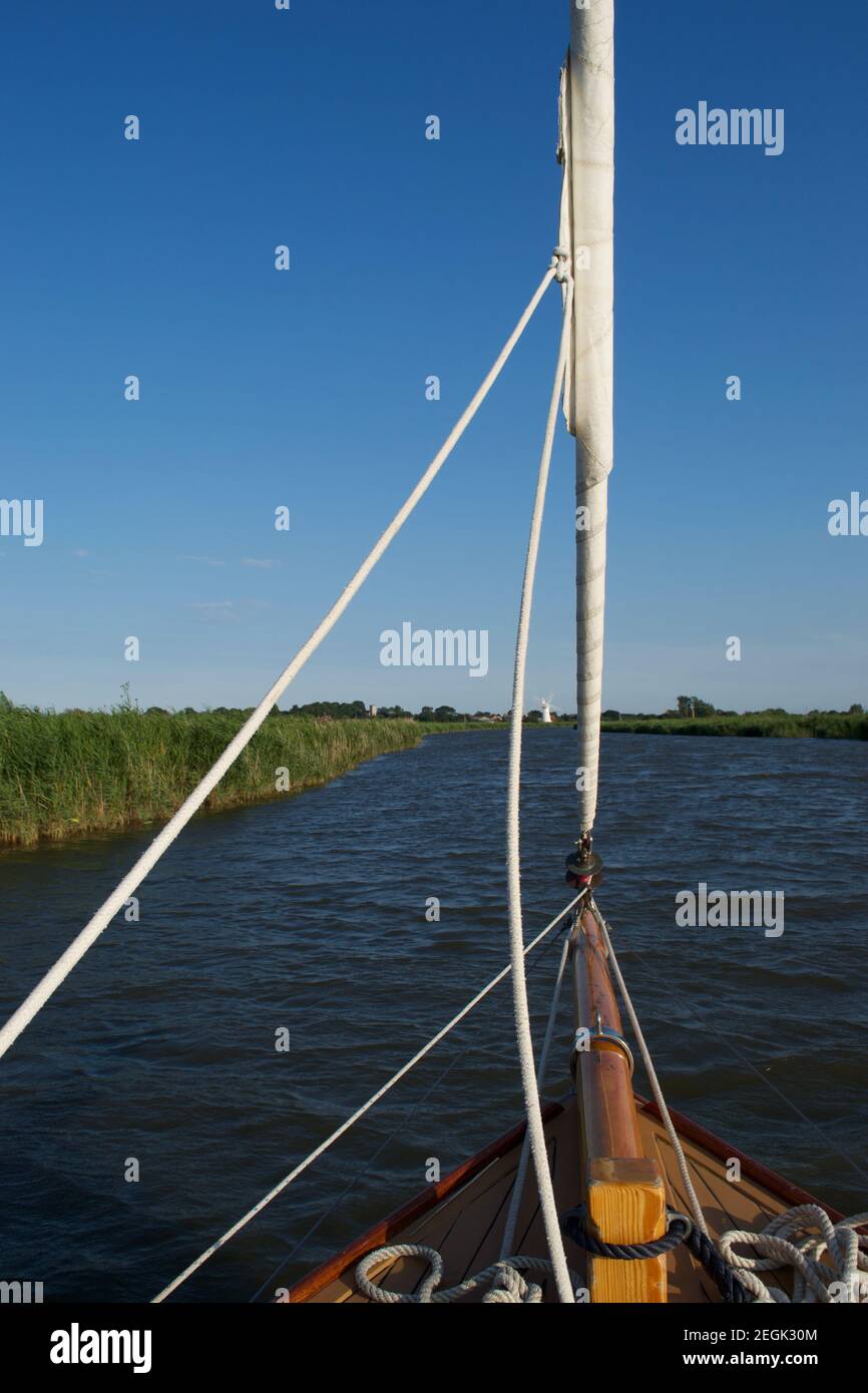 Looking up a river from the foredeck of a sailing boat (yacht). Green reedbeds on the riverbanks, and blue sky and waves ahead. Forestay, bowsprit Stock Photo