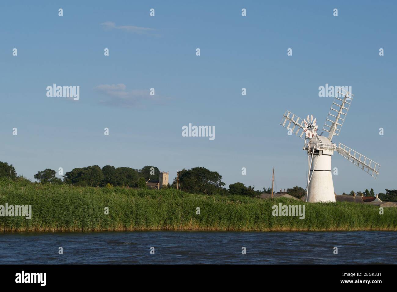 A vivid white windmill (wind pump) standing in green reedbeds on the bank of a blue river. Bright summer colours; waterside scene. Stock Photo