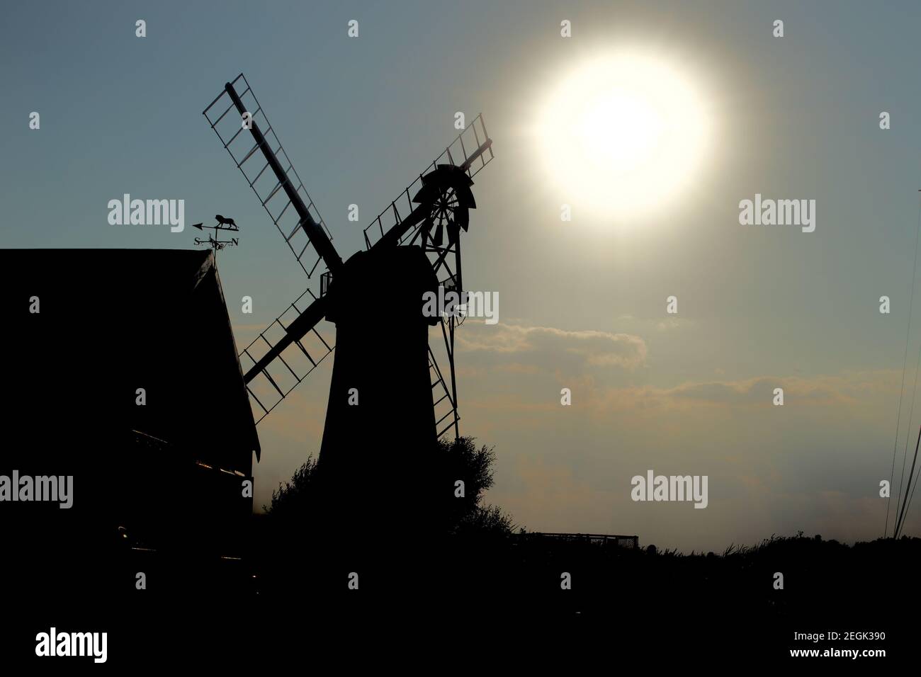 A traditional windmill (wind pump) silhouetted against a large bright low level evening sun. Shadows and darkness shroud the traditional building Stock Photo