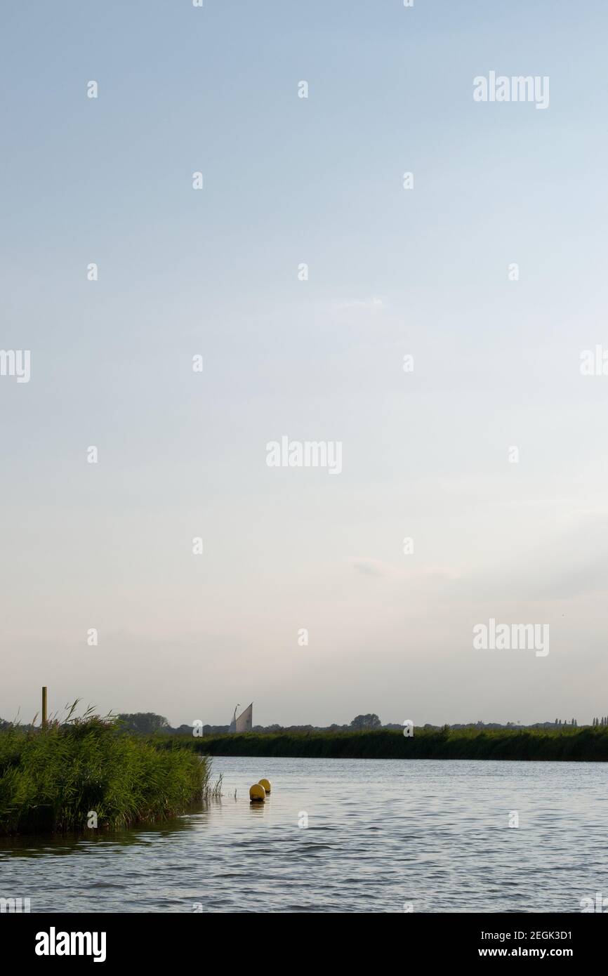 A bend in a river: buoys in a calm patch in a bend on a wide river between green reedbeds. The sail of a traditional sailing boat (yacht) in distance Stock Photo