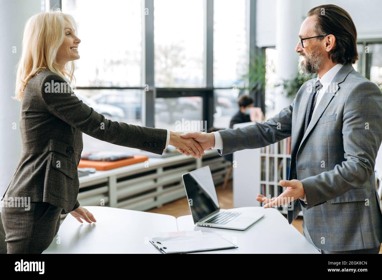 Satisfied business people are shaking hands after successful negotiations on briefing meeting. Influential colleagues in formal suits standing in modern office, coming to agreement Stock Photo