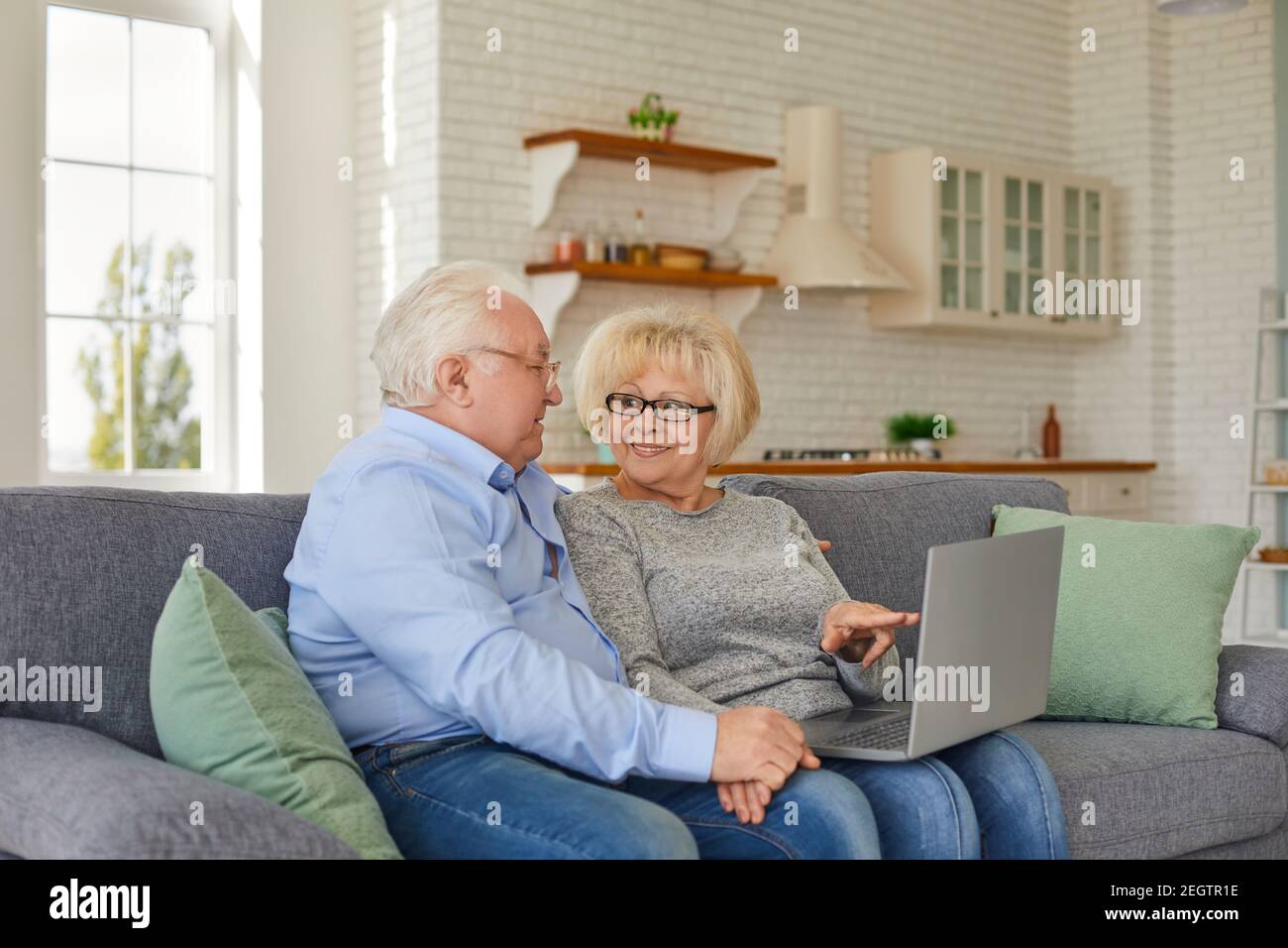Smiling mature wife and husband looking at laptop screen sitting on sofa in cozy home. Stock Photo
