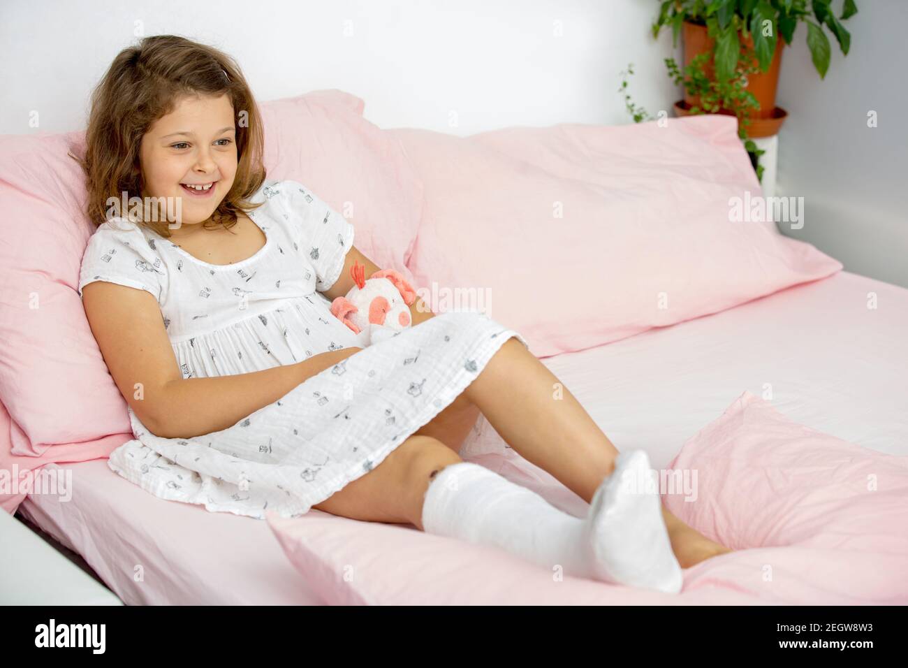 A joyful teenage girl lies on the bed in a cast on her leg, with a broken ankle. Broken leg in a child. Stock Photo