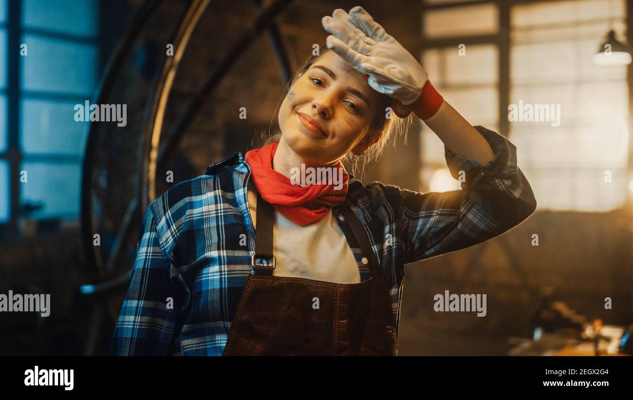 Young Beautiful Empowering Woman Rubs Her Forehead and Gently Smiles at the Camera. Authentic Fabricator Wearing Work Clothes in a Metal Workshop. Stock Photo