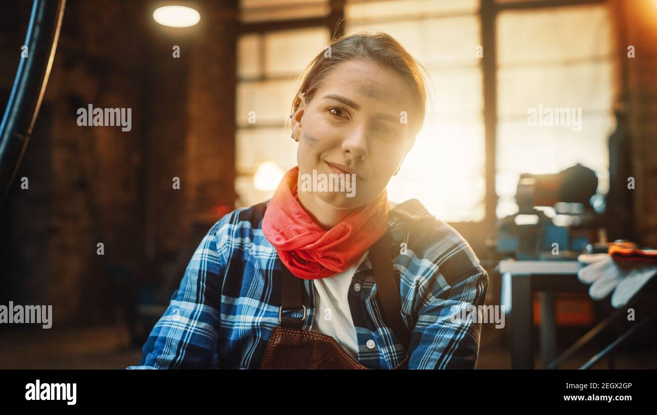 Young Beautiful Empowering Woman with Ear Piercing Gently Smiles at the Camera. Authentic Fabricator Wearing Work Clothes in a Metal Workshop. Stock Photo