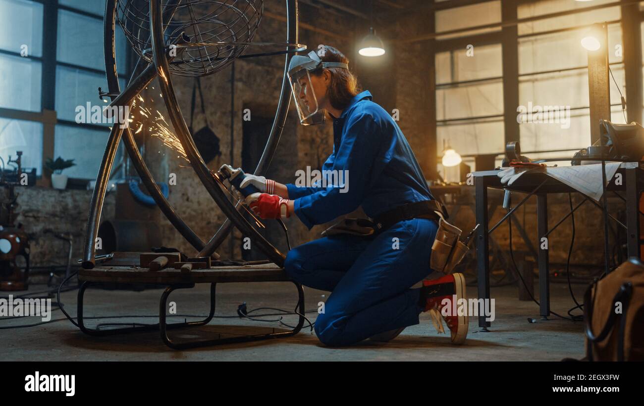 Young Contemporary Female Artist in Blue Jumpsuit is Grinding a Metal Tube Sculpture with an Angle Grinder in a Studio Workshop. Empowering Woman Stock Photo