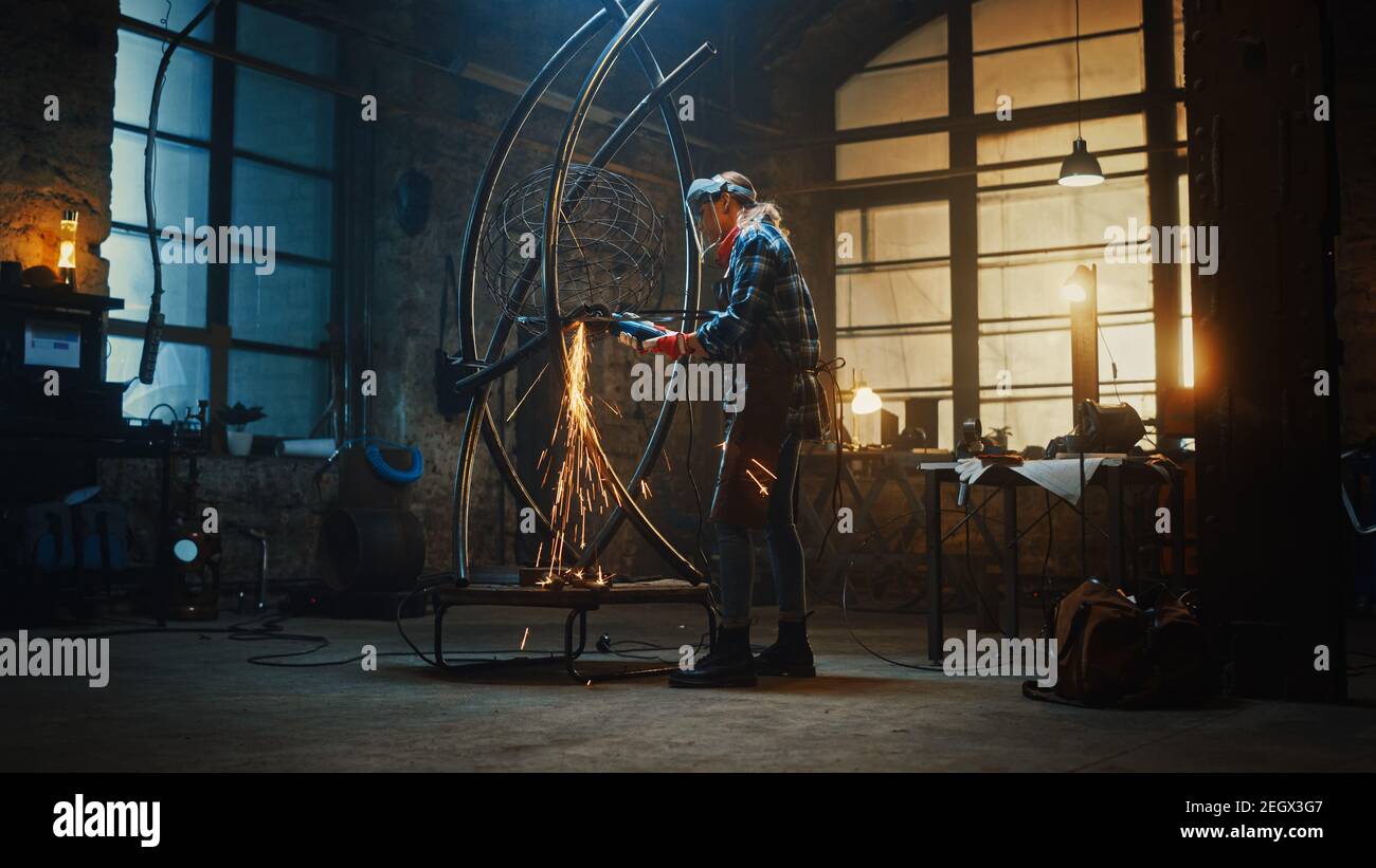 Talented Innovative Female Artist Walks Up to a Metal Tube Sculpture, Turns On and Uses an Angle Grinder in a Workshop. Contemporary Fabricator Stock Photo