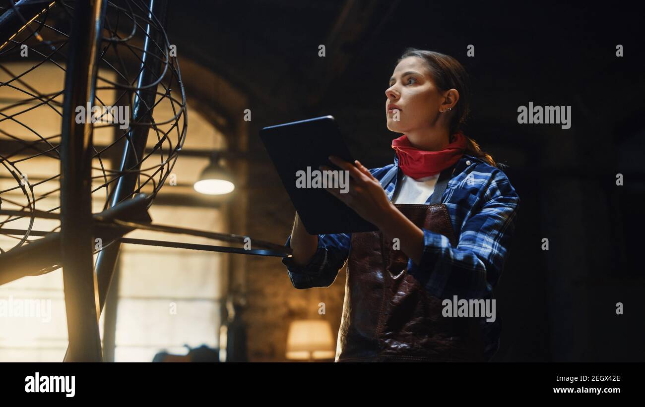 Beautiful Female Artist Sketches on a Tablet Computer Next to Brutal Metal Sculpture in Studio. Tomboy Girl Wears Checkered Shirt and Apron Stock Photo