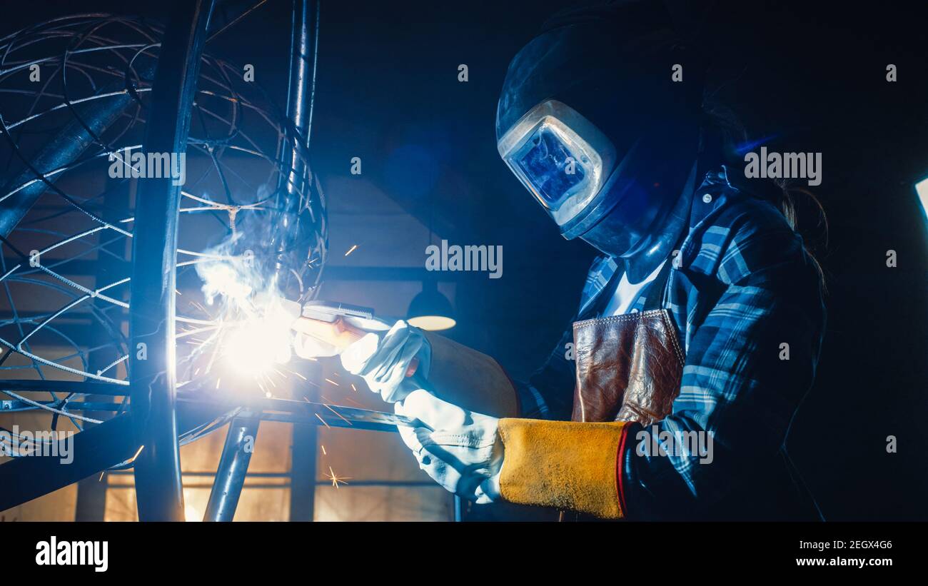 Talented Innovative Tomboy Female Artist in a Checkered Shirt is Welding an Abstract, Brutal and Expressive Metal Sculpture in a Workshop Stock Photo