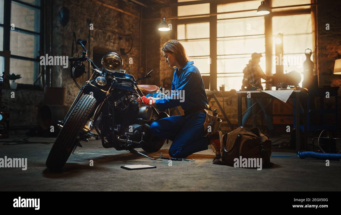 Young Beautiful Female Mechanic is Working on a Custom Bobber Motorcycle. Talented Girl Wearing a Blue Jumpsuit. She Uses a Ratchet Spanner to Tighten Stock Photo