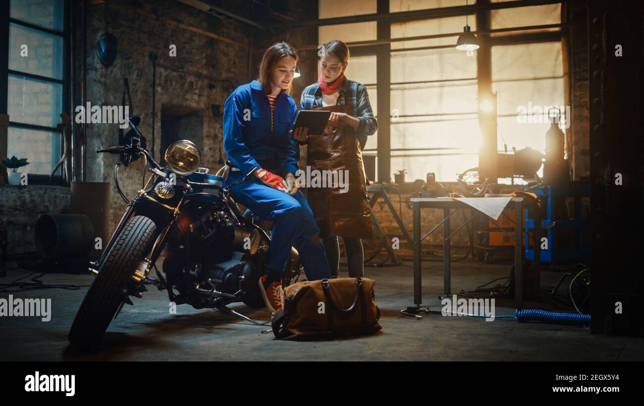 Two Young Beautiful Female are Discussing The Work Done on a Custom Bobber Motorcycle. Talented Girls Use a Tablet Computer. They are In Good Spirit Stock Photo