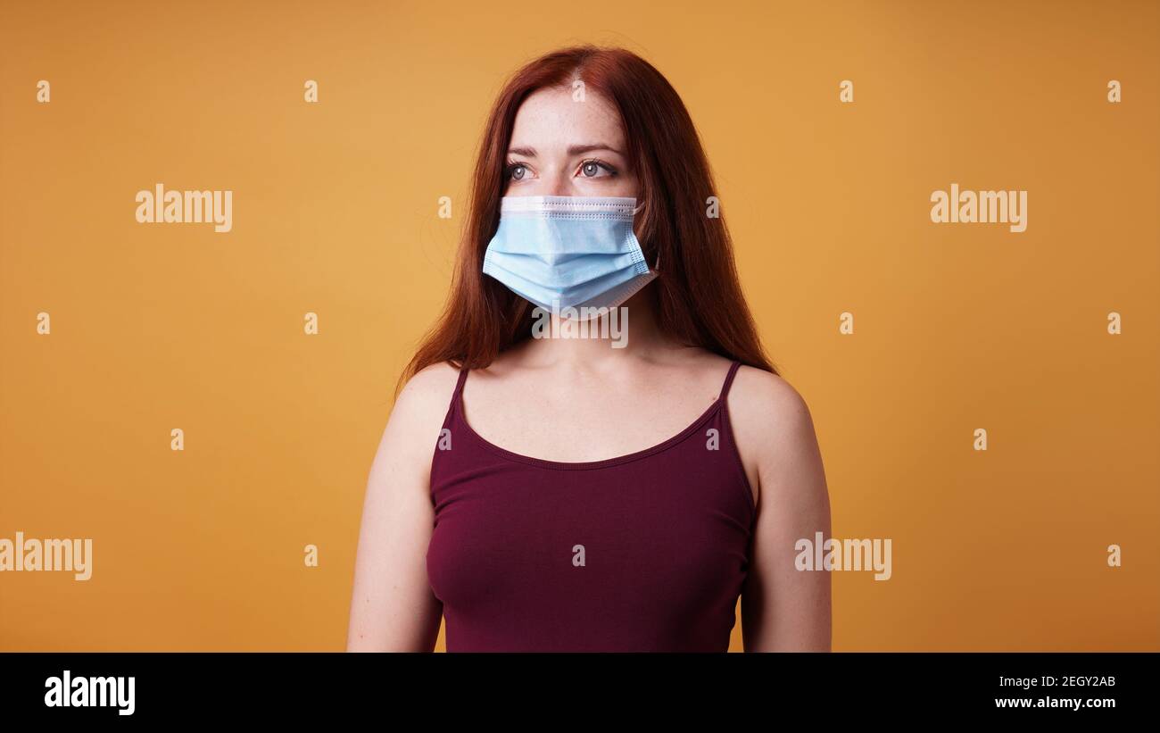 young woman wearing a medical face mask covering mouth and nose - protection against corona virus - studio portrait on orange color background with copy space Stock Photo