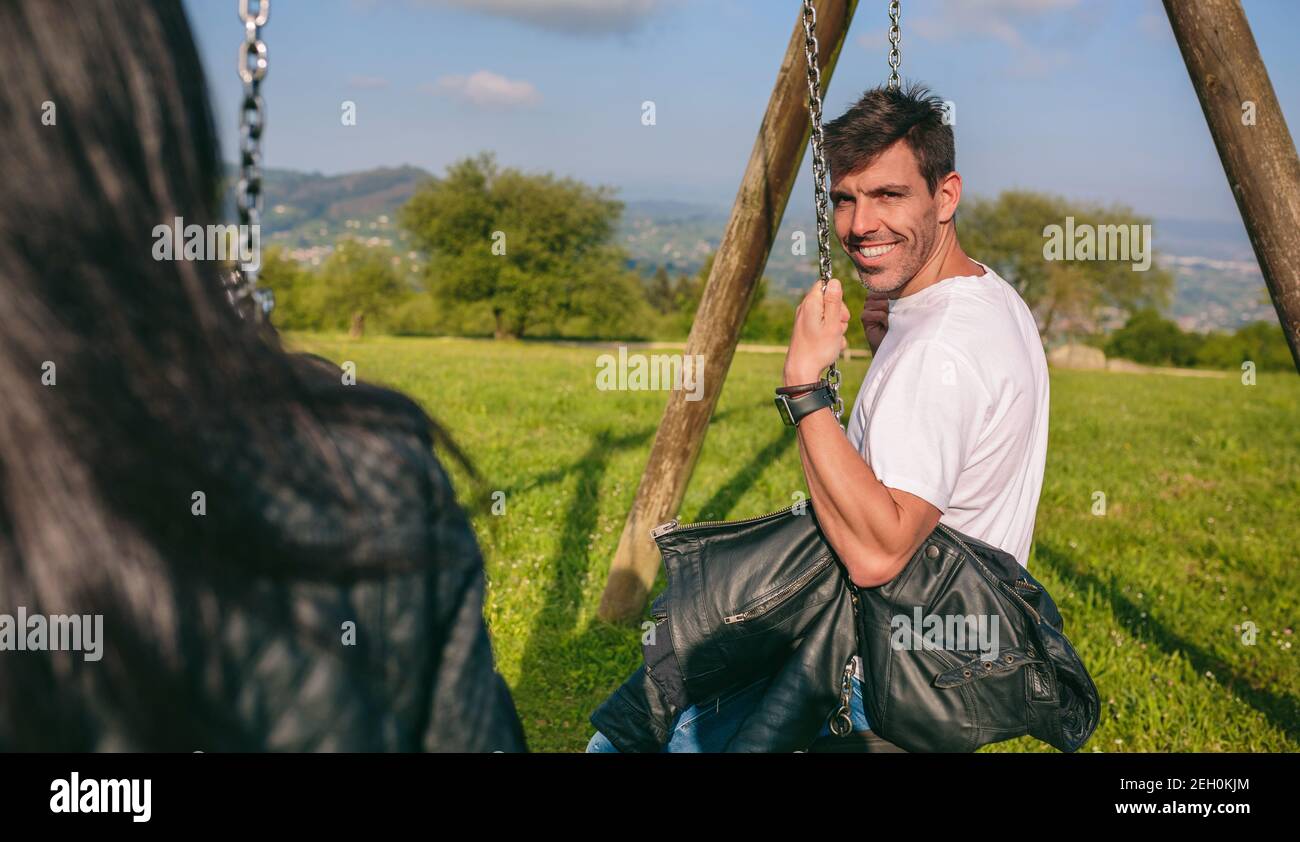 Happy young man looking camera sitting on a swing in a recreational area Stock Photo
