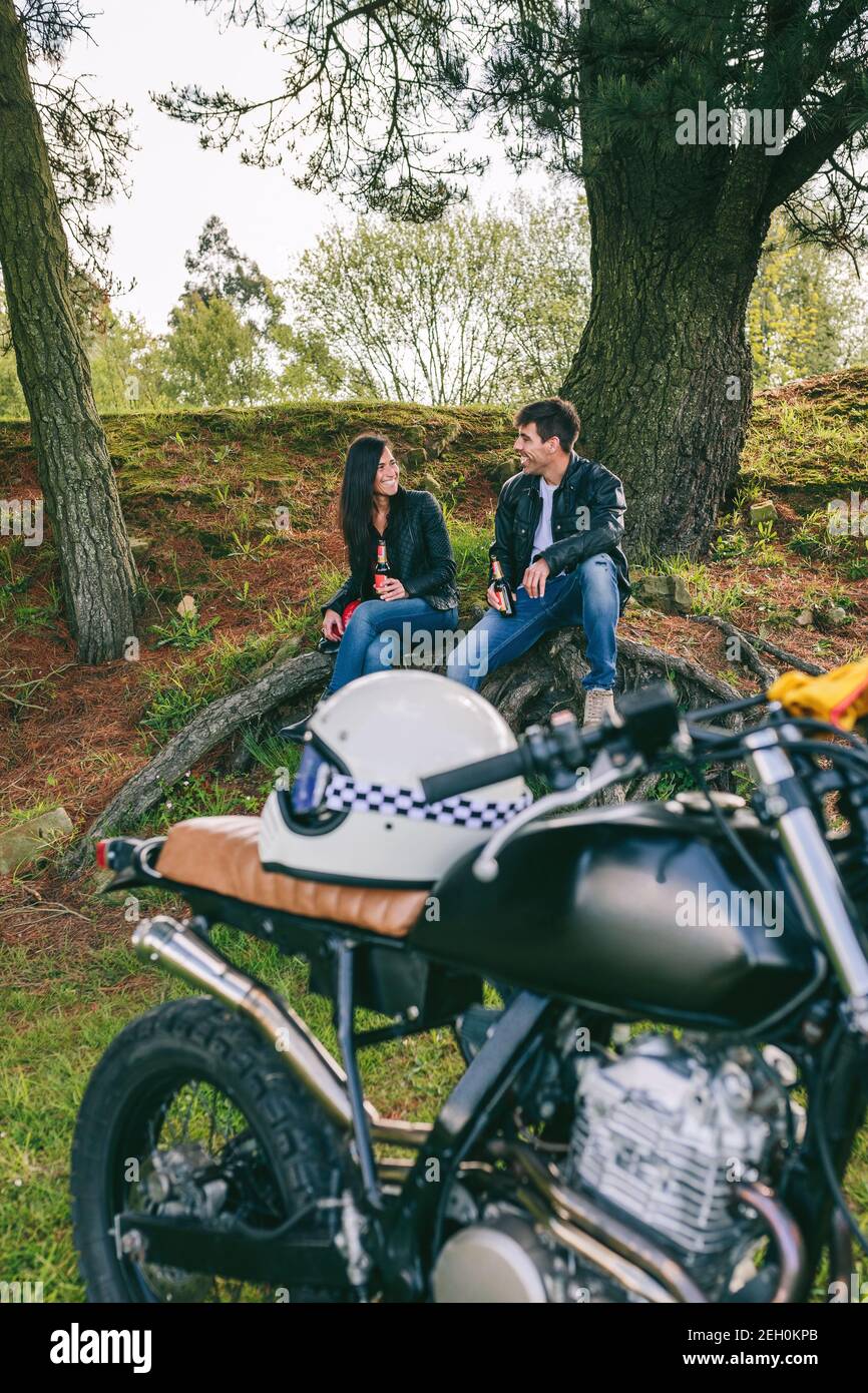 Young couple resting from a motorcycle trip having a beer outdoors with motorbike in foreground. Selective focus on couple in the background Stock Photo
