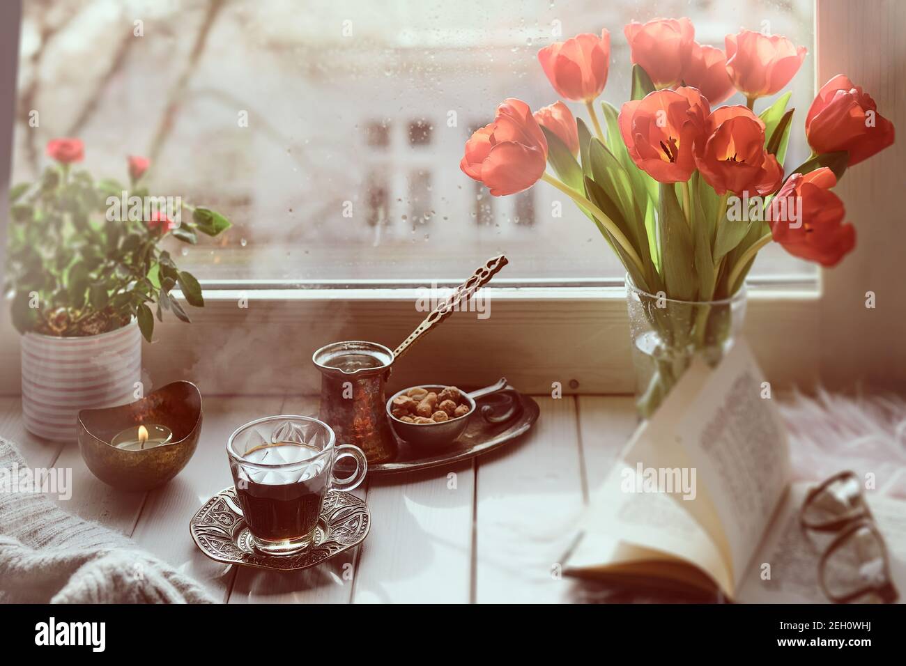 Oriental coffee in traditional Turkish copper coffee pot with flowers on window sill. Rustic wooden window sill with bunch of tulips book. Cold rainy Stock Photo