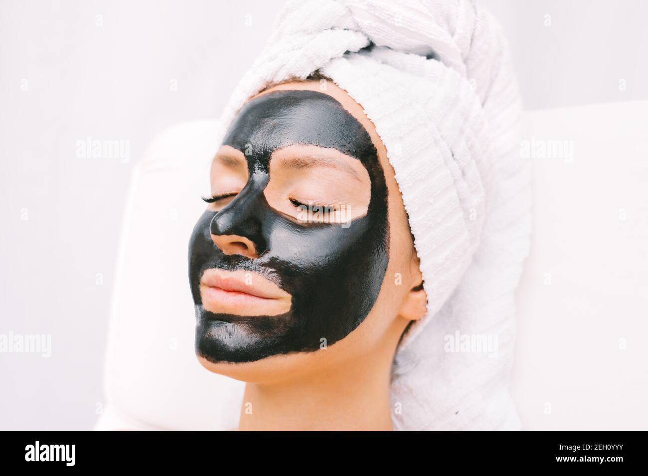 Beautiful woman with towel on her head relaxing with black purifying charcoal facial mask. Cosmetology and skincare. Beauty treatments. Spa therapy. Stock Photo