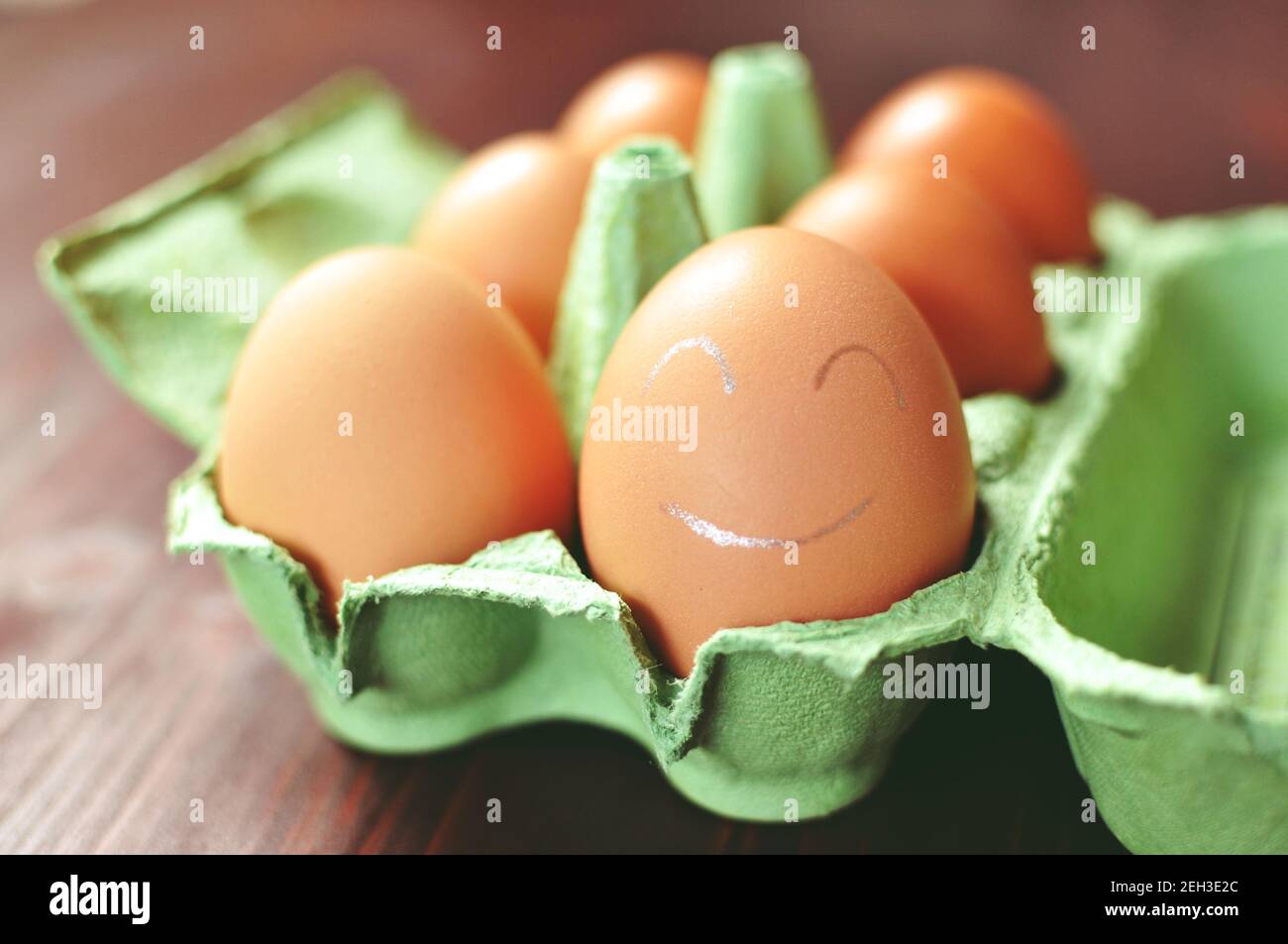 Group of brown chicken eggs in an green open form work on brown background. The closest egg is painted with white smiley face. Front view. Stock Photo