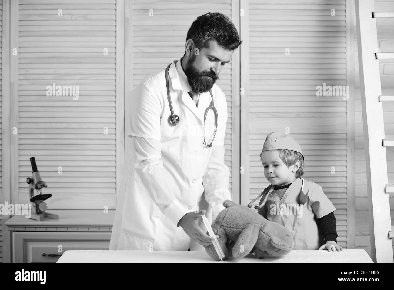 Vet and little assistant make injection to teddy bear. Father and kid with busy faces play doctor. Treatment and childhood concept. Man with beard and boy hold syringe on wooden background. Stock Photo
