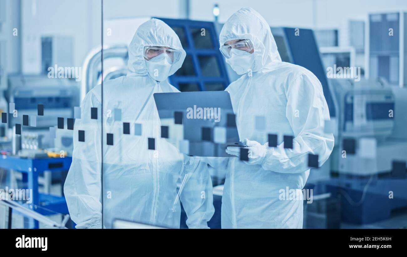 Modern Factory Sterile Workshop: Professionals in Coveralls, Masks Use Laptop and Talk. Medical Manufacturing Laboratory with High Tech CNC Machinery Stock Photo