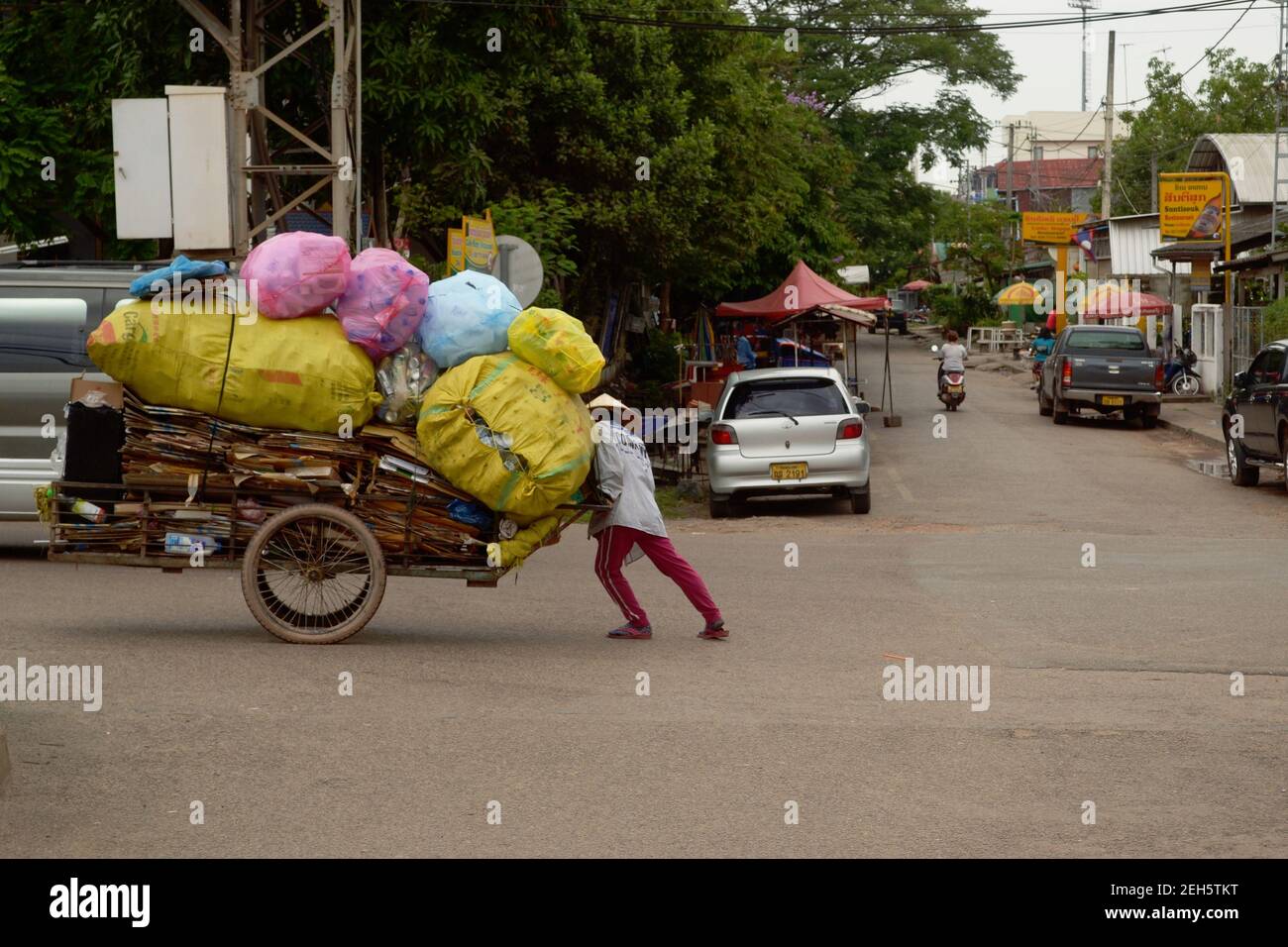 Vientiane, Laos - April, 2015: Woman is pushing heavy cart on wheels with big heap of bags with plastic bottles on a street Stock Photo