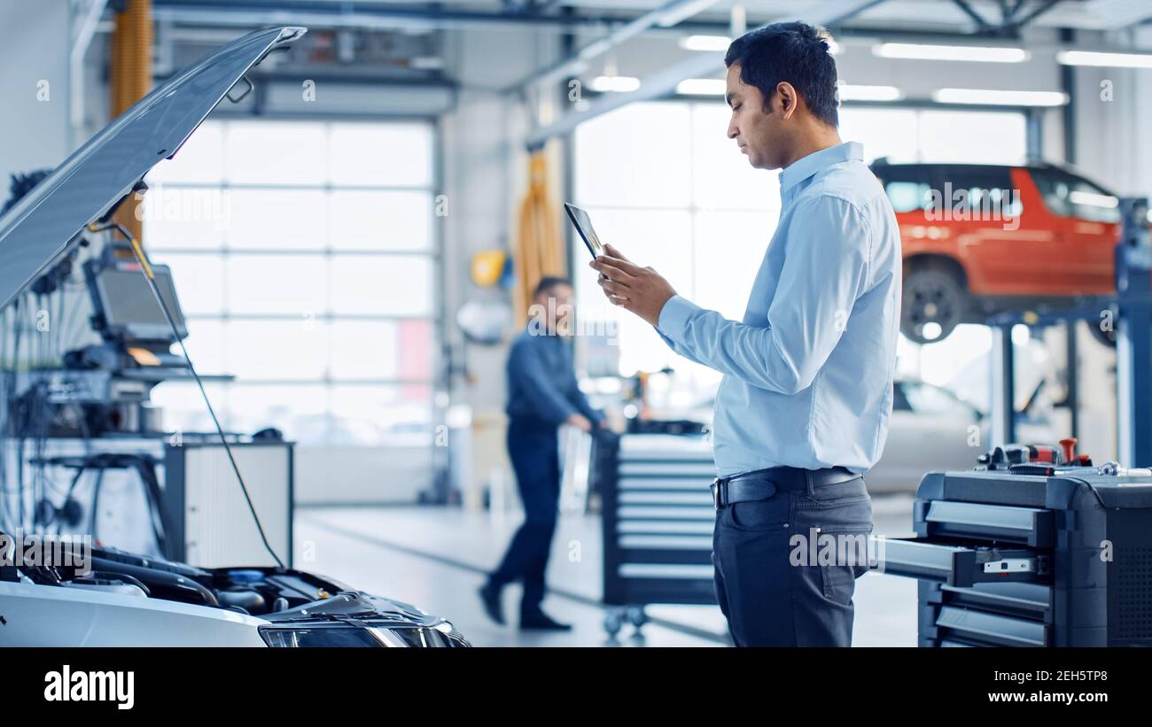 Car Service Manager Uses a Tablet Computer with an Augmented Reality Diagnostics Software. Specialist Inspecting the Car in Order to Find Broken Stock Photo