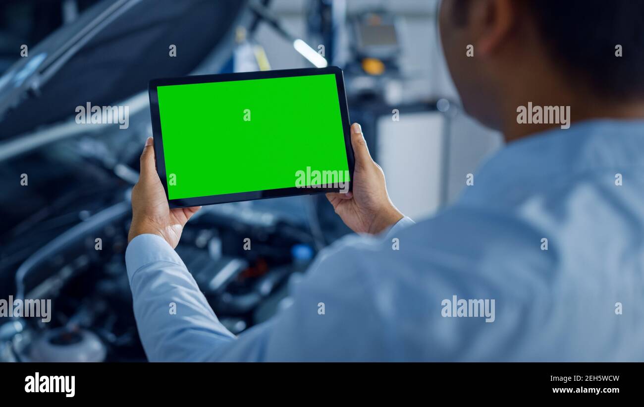 Car Service Manager or Mechanic Uses a Tablet Computer with a Green Screen Mock Up that is Pointed at an Enginer Bay. Specialist Inspecting the Stock Photo