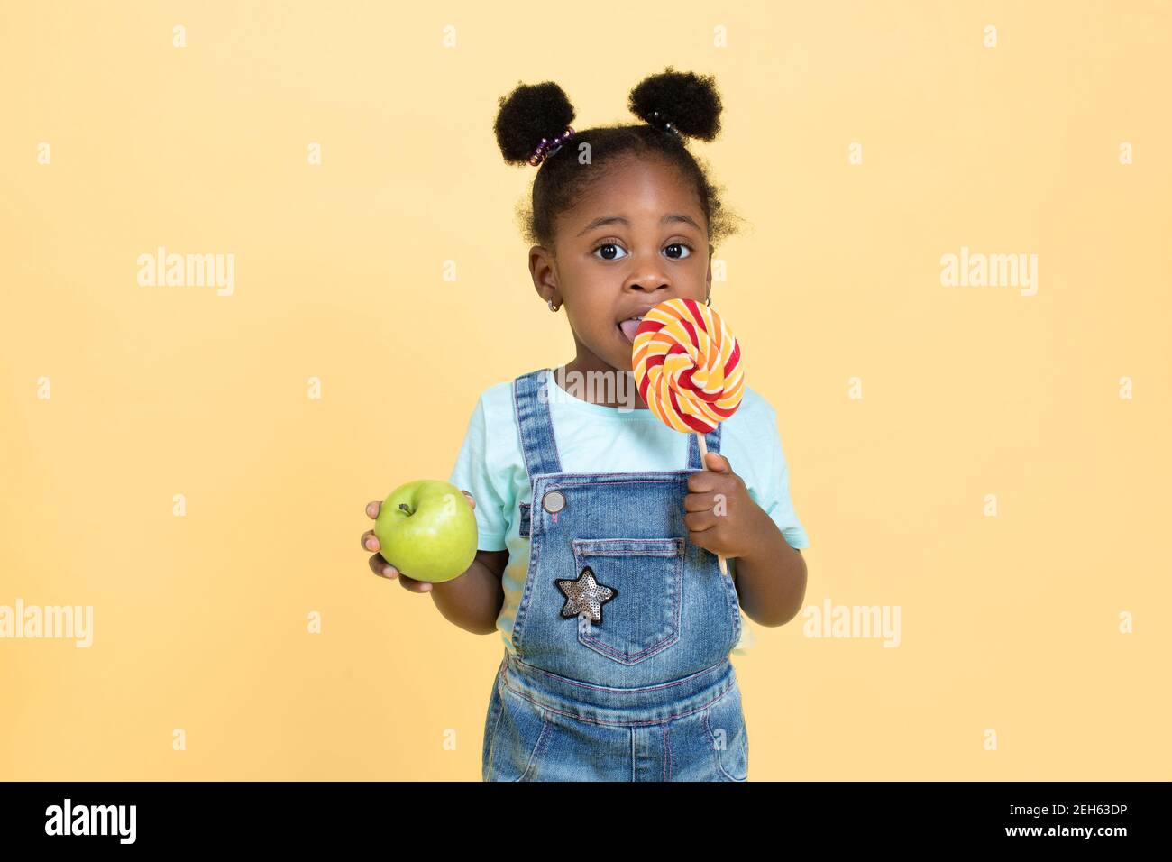 Little cute pretty African child girl eating a candy lollipop while holding  green apple in other hand, isolated on yellow background. Choice between  Stock Photo - Alamy
