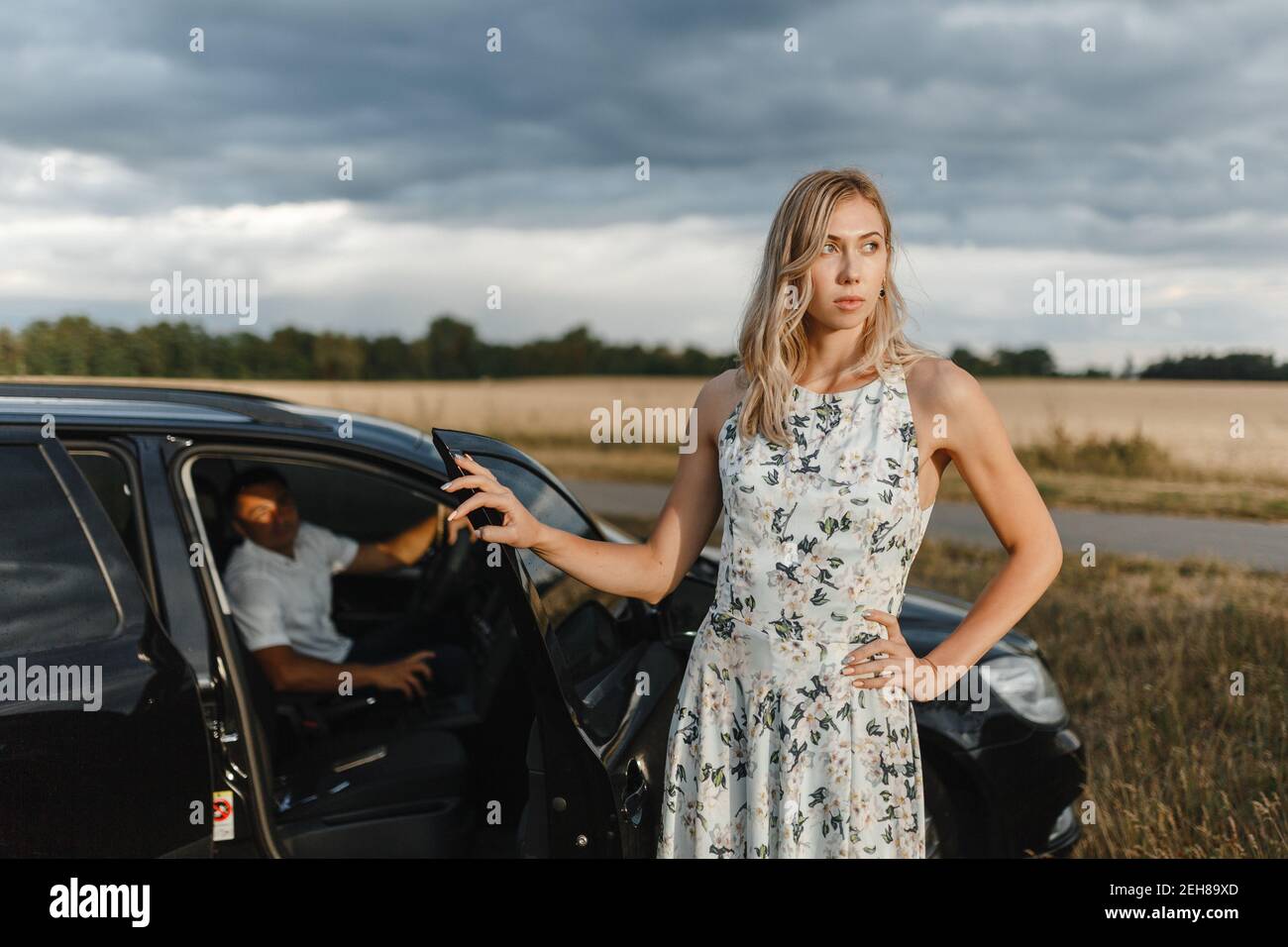 Blonde woman standing near the car door and man sitting inside the car. Couple in the car Stock Photo