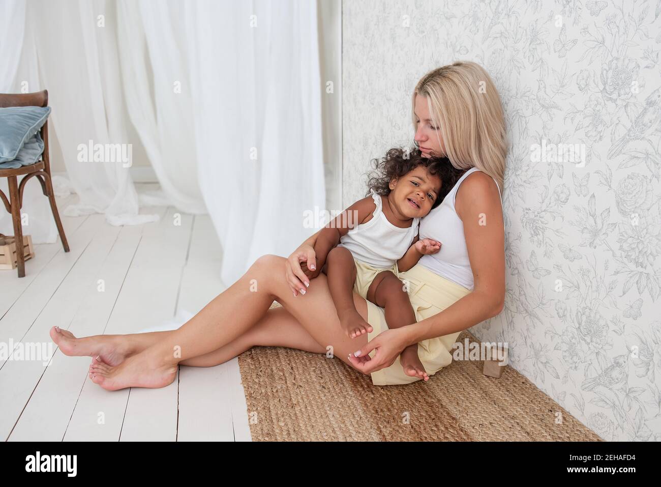 Portrait of young blonde Caucasian mother holding hugging little African American daughter in arms. Girl pressed against the womans chest. Tenderness Stock Photo