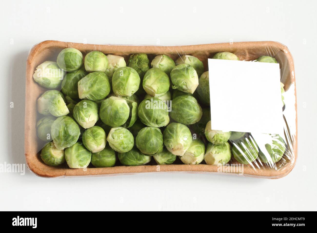 The heads of Brussels sprouts are randomly placed in a cardboard box and covered with transparent foil. The box is on a white table. Closeup. View fro Stock Photo