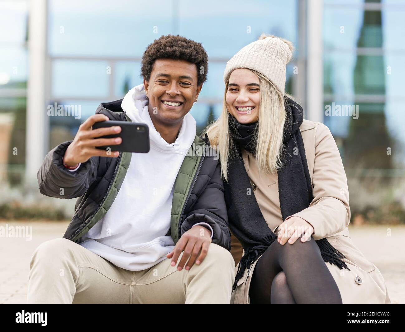 Young sincere multiethnic partners in warm clothes taking selfie phone while spending time together in town Stock Photo