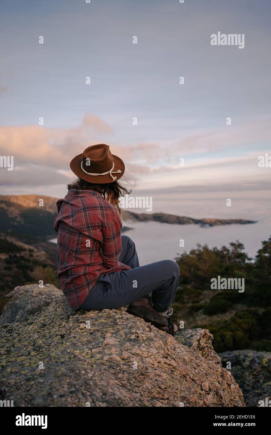 Back view of unrecognizable female tourist in hat admiring ocean with horizon from mountain under cloudy sky Stock Photo