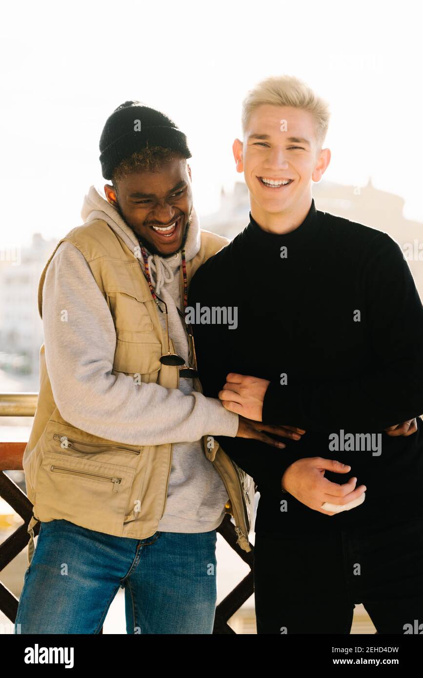 Joyful young multiracial gay couple in trendy outfits smiling and hugging while standing together on terrace on sunny day Stock Photo Alamy