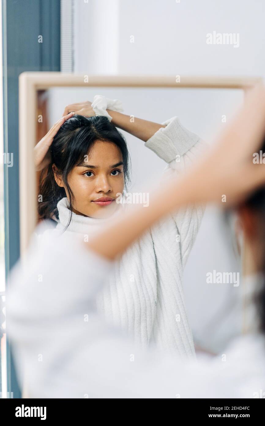 Long-haired brunette Asian Woman dressed in a white sweater, looking in the mirror and combing her hair Stock Photo