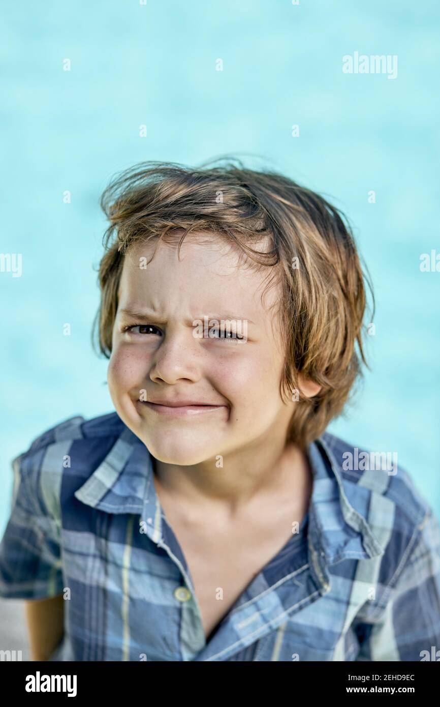 Funny little boy with blond hair in checkered shirt frowning and looking at camera against blue background Stock Photo