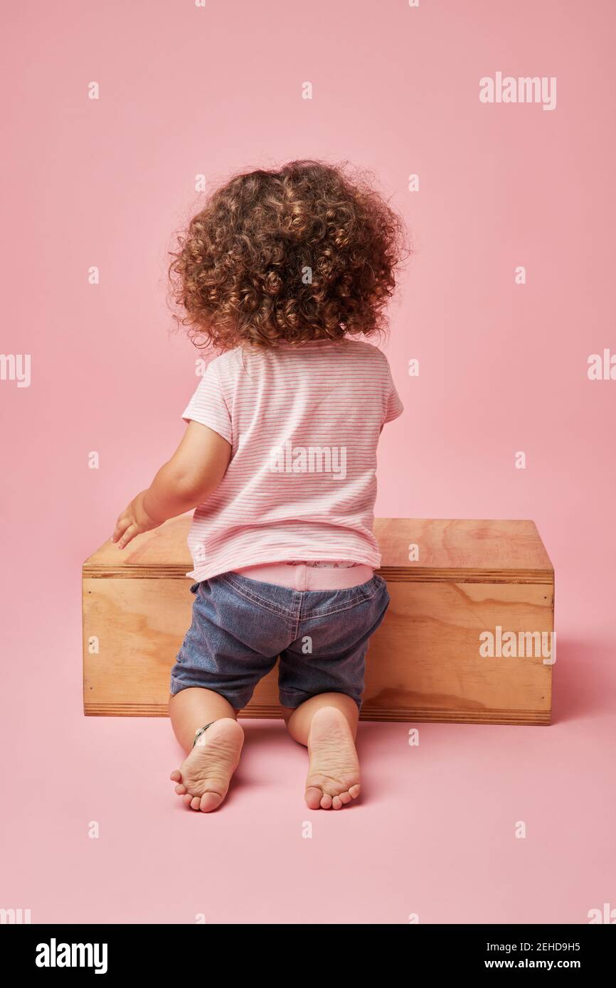 back view of unrecognizable barefoot child in t shirt and denim shorts with curly hair playing on wooden platform Stock Photo
