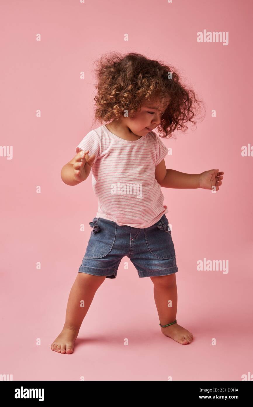 Charming barefoot child in t shirt and denim shorts with curly hair looking down while dancing on pink background Stock Photo