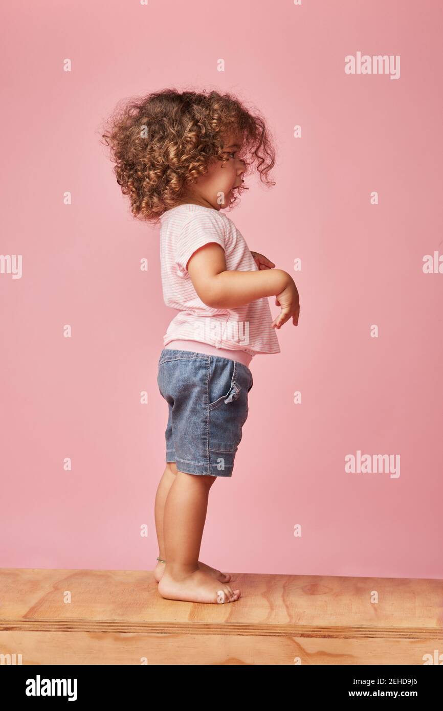 Side view of charming barefoot child in t shirt and denim shorts with curly hair looking away while standing playing on wooden platform Stock Photo