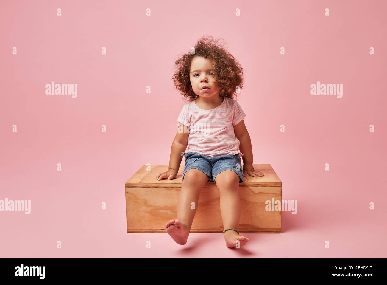 Charming barefoot child in t shirt and denim shorts with curly hair looking away while sitting on wooden platform Stock Photo