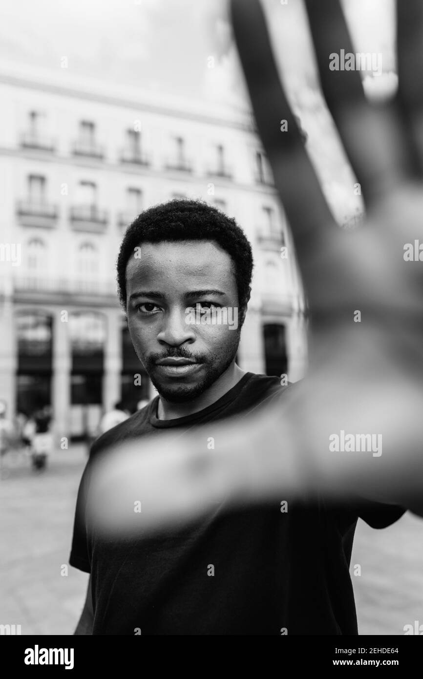 Black and white of young calm ethnic male showing stop gesture while looking at camera in town Stock Photo