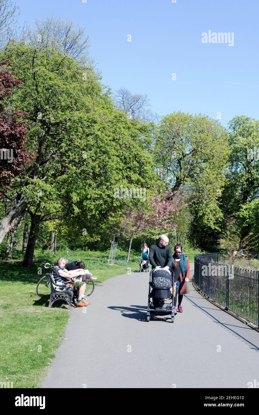 People walking along a path including couple with pushchair and lady sitting on bench in Clissold Park on a fine Spring day, Stoke Newington Stock Photo