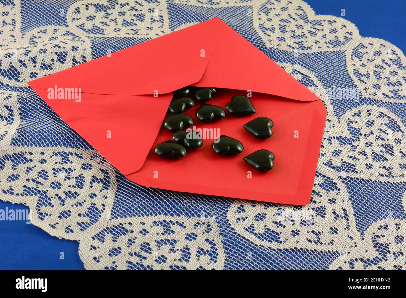 Black hearts spilling out of open red Valentine envelope on pink heart lace table runner on blue tablecloth Stock Photo