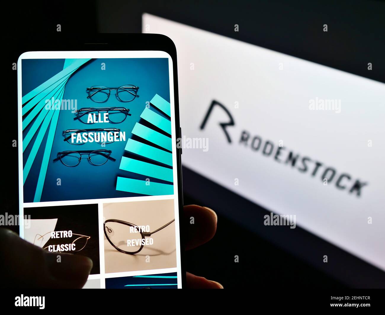 Person holding mobile phone with webpage of German eyewear manufacturer Rodenstock GmbH on screen in front of logo. Focus on center of phone display. Stock Photo