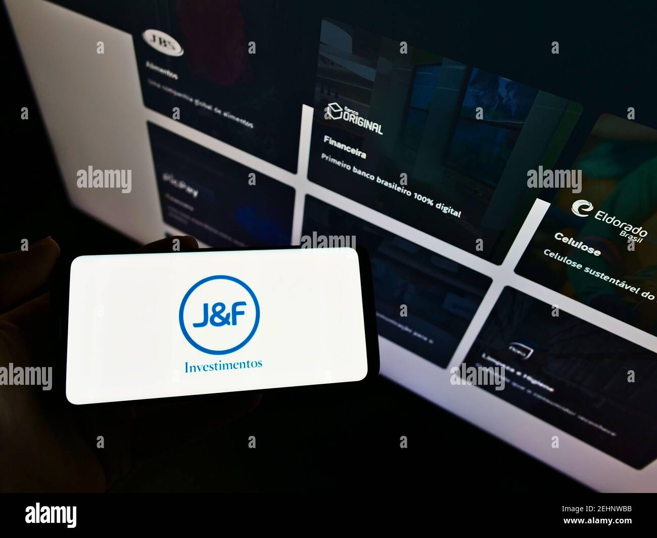 Person holding smartphone with logo of Brazilian investment company JF Investimentos S.A. on screen in front of web page. Focus on cellphone display. Stock Photo